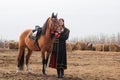 A beautiful woman with long and black hair in a historical hussar costume stands in a field with a horse Royalty Free Stock Photo