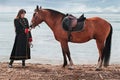 A beautiful woman with long and black hair in a historical hussar costume stands near a river with a horse Royalty Free Stock Photo