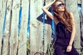 Beautiful woman with long chestnut hair wearing jumpsuit and trendy aviator sunglasses at the wooden fence