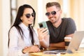 Beautiful woman and man in sunglasses look at phone and smile at round table.