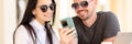 Beautiful woman and man in sunglasses look at phone and smile at round table.