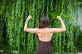 Beautiful woman with naked back over green weeping willow background. Sport girl shows back muscles. Royalty Free Stock Photo