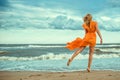 Beautiful woman in orange mini dress with flying train dancing barefoot on the wet sand at the storming sea