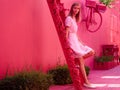 Woman in a pink dress on a bright pink street sits on a staircase decorated with flowers