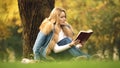 Beautiful woman reading bestseller as sitting under tree in autumn forest, hobby