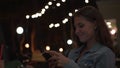 Beautiful woman reads message on background of lights of summer cafe. Media. Young woman looks at phone and smiles while