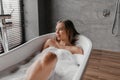 Beautiful woman relaxing in bathtub filled with foam, relaxed lady sitting in hot bath and looking at window Royalty Free Stock Photo