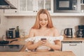 Beautiful woman sits at a table on the background of the kitchen at home. Girl with a smart phone in her hands. The blonde writes