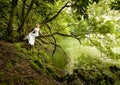 Beautiful woman sits in white long dress on branch of a tree, next to a lake