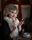 Beautiful woman sitting and correcting makeup painting lips with lipstick holding mirror in her hand at table with cup Royalty Free Stock Photo