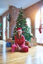 Beautiful woman sitting on the floor around christmas tree and presents wearing santa claus hat and smiling happy Royalty Free Stock Photo