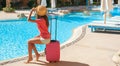 Beautiful woman sitting on pink suitcase near swimming hotel pool area. Travel, summer holidays and vacation concept