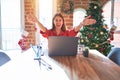 Beautiful woman sitting at the table working with laptop at home around christmas tree looking at the camera smiling with open Royalty Free Stock Photo