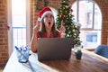 Beautiful woman sitting at the table working with laptop wearing santa claus hat at christmas amazed and surprised looking up and Royalty Free Stock Photo