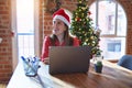 Beautiful woman sitting at the table working with laptop wearing santa claus hat at christmas looking away to side with smile on Royalty Free Stock Photo