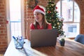 Beautiful woman sitting at the table working with laptop wearing santa claus hat at christmas smiling with happy face looking and Royalty Free Stock Photo