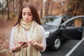 Beautiful woman smiling while stands near the car. Girl is using a smartphone. Autumn concept. Autumn forest journey by