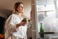 Beautiful woman standing on kitchen at night and looking in open fridge holding green apple in hand Royalty Free Stock Photo