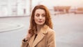 Beautiful woman standing outdoors posing for camera in an autumn beige coat, beautiful hair with urban city background Royalty Free Stock Photo