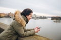 Beautiful woman standing by river in city Prague, text messaging. girl looking at phone on riverbank, with Prague bridge