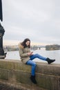 Beautiful woman standing by river in city Prague, text messaging. girl looking at phone on riverbank, with Prague bridge