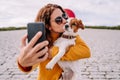 A beautiful woman staying in a park with her cute little dog. She is taking a self-portrait photo with the phone while kissing her