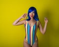 Beautiful woman in a striped swimsuit in a blue wig posing on a yellow background. Portrait of a glamorous girl. Royalty Free Stock Photo