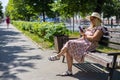 A beautiful woman in a sundress sits on a bench in a city park in summer. A woman in a dress writes a message on her