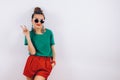 Beautiful woman in sunglasses wearing in red shorts and green T-shirt standing near white wall, smiling and shows Royalty Free Stock Photo