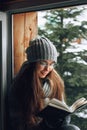 Beautiful woman in a sweater  sitting home by the window. Blurred winter snow tree background. Morning, coziness, winter and peopl Royalty Free Stock Photo