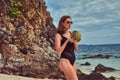 Beautiful woman in a swimsuit, holds coconut with a tube, standing on a beach near big reef stones, enjoys a vacation on Royalty Free Stock Photo