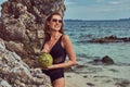 Beautiful woman in a swimsuit, holds coconut with a tube, standing on a beach near big reef stones, enjoys a vacation on Royalty Free Stock Photo