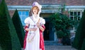 Beautiful woman in vintage Georgian dress and bonnet standing in garden of stately home