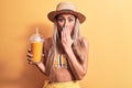 Beautiful woman wearing bikini and hat drinking healthy orange juice over yellow background covering mouth with hand, shocked and Royalty Free Stock Photo