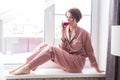 Beautiful woman wears pajamas with a glass of red wine sits on the windowsill near the beautiful window at home. Stay Royalty Free Stock Photo