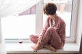 Beautiful woman wears pajamas with a glass of red wine sits on the windowsill near the beautiful window at home. Stay Royalty Free Stock Photo