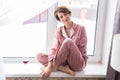 Beautiful woman wears pajamas with a glass of red wine sits on the windowsill near the beautiful window at home. Stay Royalty Free Stock Photo