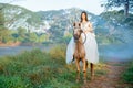 Beautiful woman with white dress stay on brown horse on riding through the road way with background of fog or smoke during early Royalty Free Stock Photo