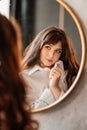 a beautiful woman in a white shirt looks at her reflection in a round mirror.