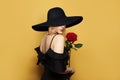 Beautiful woman in wide black broad brim hat with blonde hair and makeup holding red rose flower on yellow background Royalty Free Stock Photo