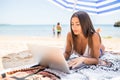 Beautiful woman working online on laptop while lying on beach under sun umbrella near sea. Happy smiling freelancer girl relaxing