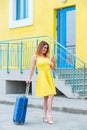 A beautiful woman in a yellow dress with a blue suitcase uses a mobile. A girl calls a taxi using an application on a Royalty Free Stock Photo