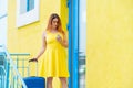 A beautiful woman in a yellow dress with a blue suitcase uses a mobile. A girl calls a taxi using an application on a Royalty Free Stock Photo