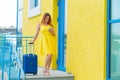 A beautiful woman in a yellow dress with a blue suitcase uses a mobile. A girl calls a taxi using an application on a Royalty Free Stock Photo