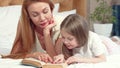 Beautiful woman reading a book with her cute little daughter, lying on bed at home