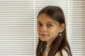 A beautiful 11-year-old girl against the background of window blinds, close-up, selective focus. Concept: waiting for parents, orp