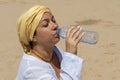 A beautiful 40-year-old woman drinks water from a plastic bottle in the desert against the background of sand. Concept: thirst and