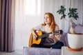 Beautiful young adult girl with long red hair playing the guitar sitting at home in light interior, selective focus. Thoughtful