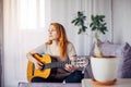 Beautiful young adult girl with long red hair playing the guitar sitting at home in light interior, selective focus. Thoughtful