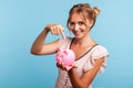 Beautiful young adult woman in summer dress holding piggy bank and pointing with index finger, suggesting you to put coins inside Royalty Free Stock Photo